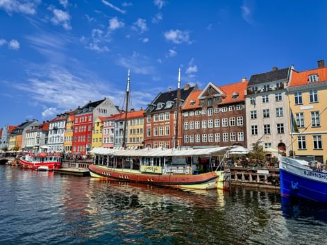 Historic Nyhavn harbor in Copenhagen with colorful townhouses, boats moored along the canal, and outdoor cafes under white awnings on a sunny day.