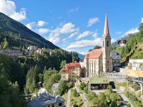 Church with distinctive red spire in Bad Gastein, surrounded by colorful buildings on a hillside with forested mountains and blue sky.