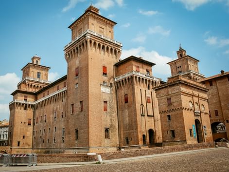 Castello Estense in Ferrara mit vier Ecktürmen und zinnenbesetzten Mauern aus braunem Backstein unter blauem Himmel mit weißen Wolken.