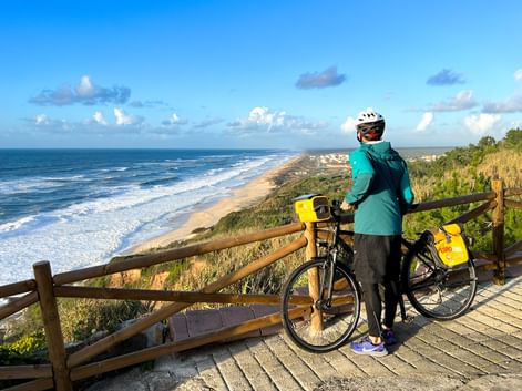 Radfahrer mit Helm und türkisfarbener Jacke steht mit Fahrrad auf hölzernem Aussichtspunkt mit Blick auf langen Sandstrand und Atlantik bei Coimbra.