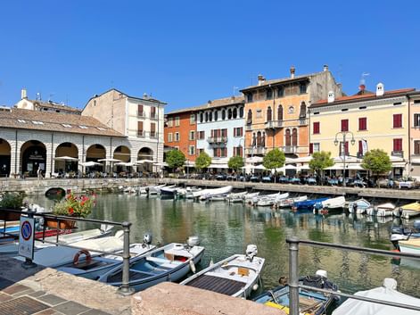 Harbor of Desenzano at Lake Garda with moored boats, colorful historic buildings, and white arched colonnade under clear blue sky.