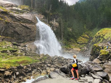 Cyclist with yellow backpack viewing Krimml Waterfalls cascading over rocky cliffs surrounded by green forest and moss-covered rocks.