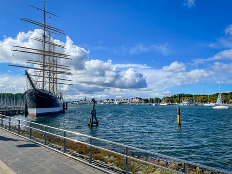 Large four-masted sailing ship moored at Travemünde harbor entrance with blue water, white clouds, and coastal town in background.
