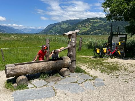 Cyclist in red with child at rustic wooden fountain in Schüttdorfer Moor. Touring bike with yellow panniers parked nearby, mountains in background.