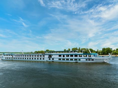 White river cruise ship MS Switzerland sailing on calm water with blue sky. The multi-deck vessel features rows of windows and a sun deck.