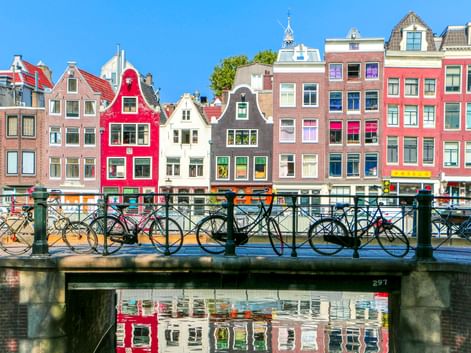 Reihe bunter historischer Grachtenhäuser in Amsterdam, die sich im Wasser spiegeln. Fahrräder stehen am Brückengeländer im Vordergrund.