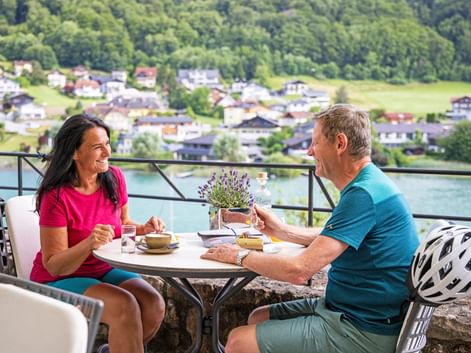 Zwei Radfahrer genießen Kaffee an einem Terrassentisch mit Blick auf den Mattsee, mit Dorf und bewaldeten Hügeln im Hintergrund.