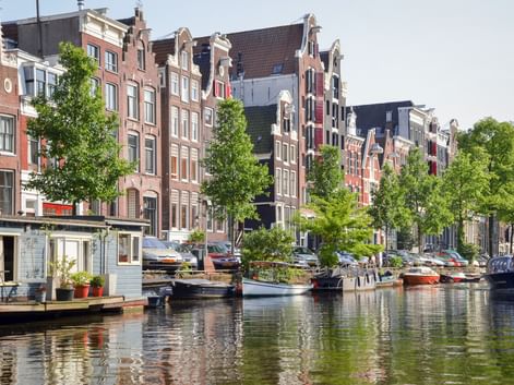 Amsterdamer Gracht gesäumt von traditionellen niederländischen Giebelhäusern und grünen Bäumen. Mehrere Hausboote liegen am Ufer.