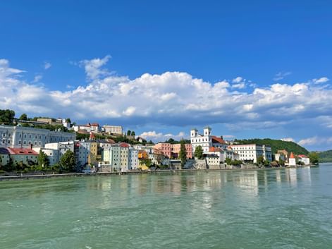 Bunte Gebäude von Passau entlang der Donau mit sichtbarer weißer Kirche und Türmen. Blauer Himmel mit weißen Wolken über der Hangstadt.