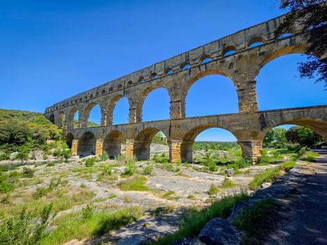 The ancient Roman Pont du Gard aqueduct with three tiers of arches spanning across a dry riverbed in Provence under a clear blue sky.