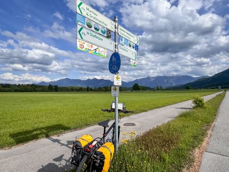 Bicycle with yellow panniers at directional signpost in Murnauer Moos. Green fields and mountains under cloudy sky in background.