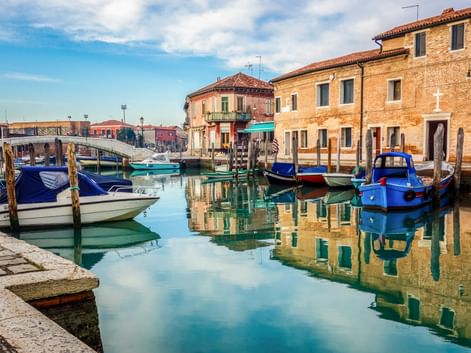 Bunte Boote am ruhigen Kanal in Murano mit traditionellen Backsteingebäuden, die sich im Wasser spiegeln, unter blauem Himmel.