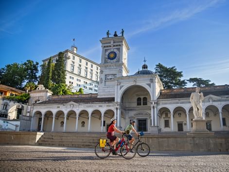 Two cyclists riding past the historic clock tower and colonnade at Piazza della Libertà in Udine, Friuli, under a blue sky.