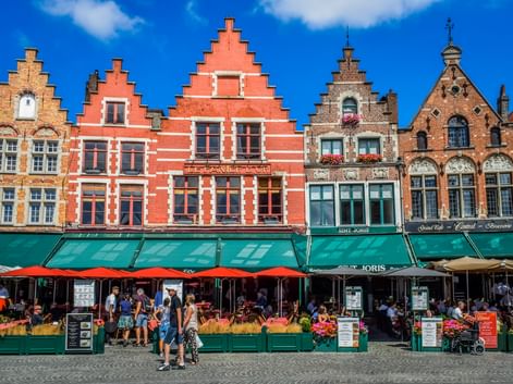 Row of traditional Flemish gabled houses with red, beige, and brown facades at Bruges Grote Markt. Green awnings shade outdoor cafes with red umbrellas.