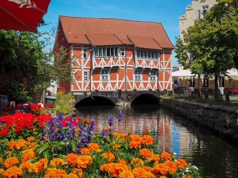 Orange half-timbered house with red tile roof spanning a canal in Wismar. Colorful flowers bloom in the foreground along the waterway.