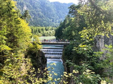 Lechfall waterfall near Füssen with turquoise water cascading over stepped weir. Visitors on footbridge, surrounded by lush forest and mountains.