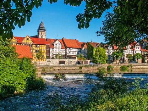 View of Hann. Münden old town with colorful half-timbered houses, red roofs, and church tower. River with weir in foreground, framed by green trees.