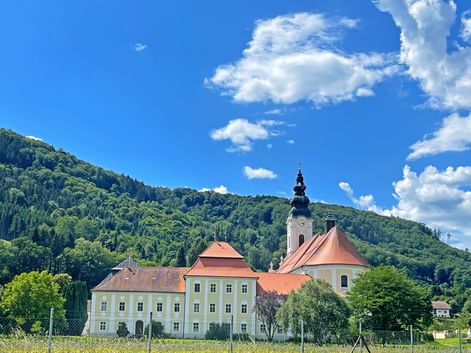 Stift Engelszell monastery with yellow buildings, red roofs, and church tower against forested hills under blue sky with white clouds.