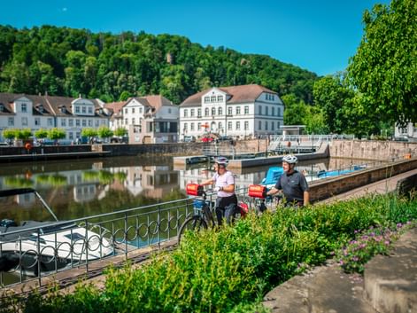 Zwei Radfahrer mit Tourenrädern am Barockhafen in Bad Karlshafen. Weiße historische Gebäude säumen das Ufer mit bewaldeten Hügeln dahinter.