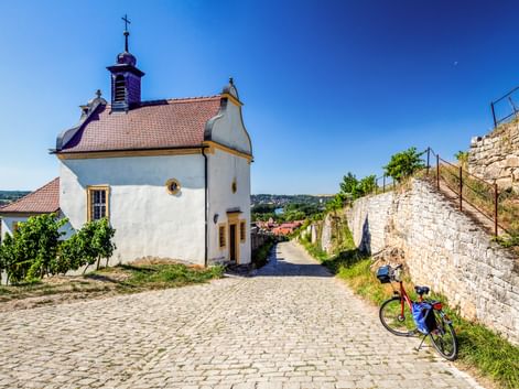 Weiße Kapelle mit orangefarbenem Dach und Glockenturm neben einem Kopfsteinpflasterweg. Ein Reiserad mit blauen Taschen steht an einer Steinmauer.