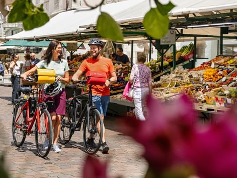 Two cyclists with e-bikes at a farmers market in Bozen, South Tyrol. Colorful fruit and vegetable stalls line the street under white awnings.