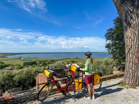Female cyclist with touring bike and yellow panniers overlooking Lake Massaciuccoli. Wide lake view with green vegetation and blue sky.