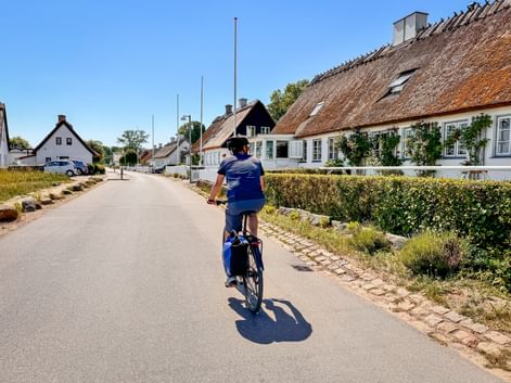 Cyclist riding through a quiet Danish village street on Zealand with traditional thatched-roof houses and hedges under a clear blue sky.