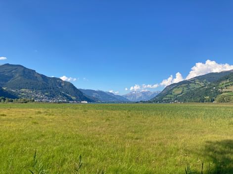 Grüne Wiese mit Blick auf Bergtal bei Zell am See. Alpengipfel und Dörfer in der Ferne unter blauem Himmel mit weißen Wolken.