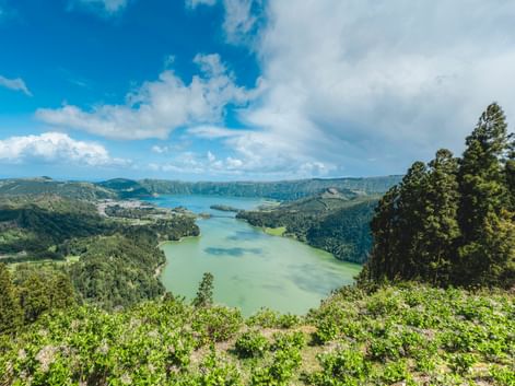 Panoramablick auf einen Vulkankratersee auf den Azoren mit türkis-grünem Wasser, umgeben von bewaldeten Hügeln unter blauem Himmel.