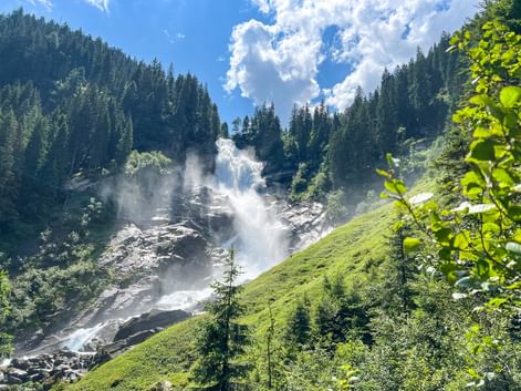 Upper Krimml Waterfalls cascading down rocky slopes with mist, surrounded by green alpine meadows and dense conifer forests under blue sky.
