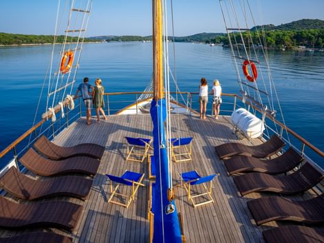 Sun deck of MS Macek with brown loungers and blue chairs. Passengers stand at the railing viewing the calm blue water and green hills.