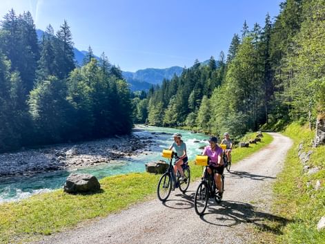 Drei Radfahrer auf einem Weg neben der türkisfarbenen Saalach bei Lofer, umgeben von dichten Wäldern und Bergen unter blauem Himmel.