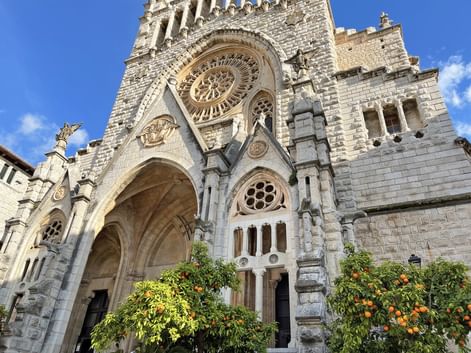 Gotische Kathedralenfassade in Sóller mit Rosettenfenster, Spitzbogeneingang und Orangenbäumen im Vordergrund vor blauem Himmel.