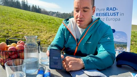 Mann in türkisfarbener Jacke an Tisch im Freien mit Handy, Wasser und Obstkorb. Eurobike-Banner hinter ihm in grüner Alpenlandschaft.