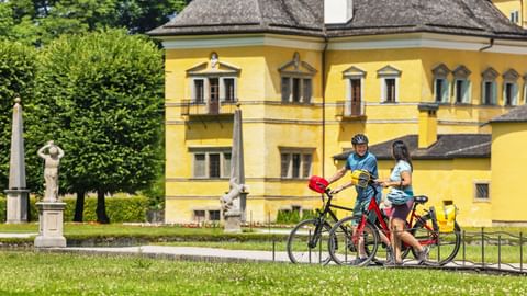 Drei Radfahrer machen Pause mit ihren Rädern vor dem gelben Schloss Hellbrunn in Salzburg, umgeben von grünen Rasenflächen und Statuen.