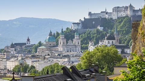 Panoramablick auf Salzburgs Altstadt mit Festung Hohensalzburg auf dem Hügel, barockem Dom mit grünen Kuppeln und Bergen im Hintergrund.