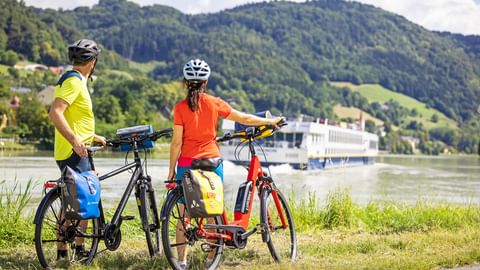 Radler am Donauradweg blicken auf ein vorbeifahrendes Schiff