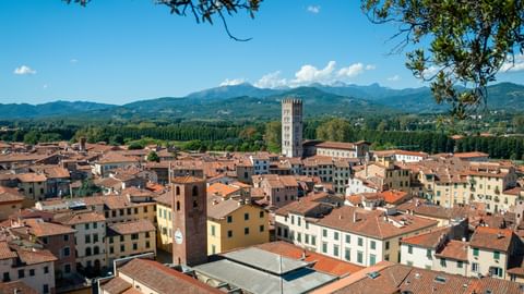 Aerial view of Lucca's historic center with terracotta rooftops, white bell tower, and mountains in the background under blue sky.