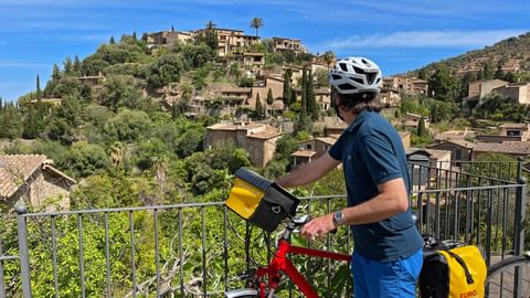 Radfahrer mit rotem Fahrrad und gelben Taschen an einem Aussichtspunkt mit Blick auf das Bergdorf Deia auf Mallorca unter blauem Himmel.