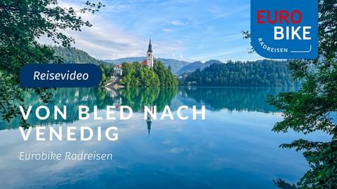 Bleder See in Slowenien mit kleiner Insel mit Kirche mit rotem Dach und Glockenturm, umgeben von bewaldeten Bergen und blauem Himmel.