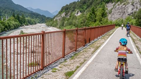 Child with blue helmet cycling on Alpe Adria bike path alongside river in Kanaltal valley, with mountains and red railing visible.