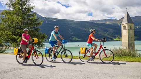 Three cyclists with yellow panniers riding along Lake Reschen. The famous church tower rises from the turquoise water, with mountains in the background.