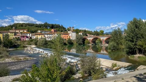 Steinbrücke mit Bögen über einem Fluss in der Toskana. Bunte Gebäude säumen den Hügel hinter der Brücke, mit Bäumen und Flussbett im Vordergrund.