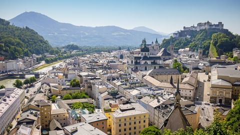 Aerial view of Salzburg's old town with Hohensalzburg Fortress on a hill, historic buildings, church domes, and mountains in the background.