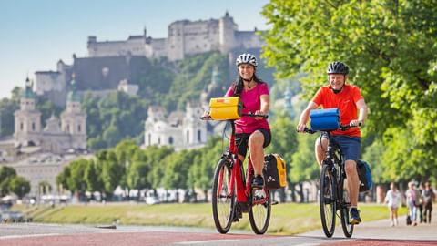 Two cyclists with panniers riding on red cycle path along the Salzach River in Salzburg with Hohensalzburg Fortress visible on the hill.