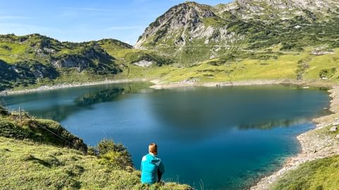 Person in türkisem Shirt sitzt auf grasigem Hang mit Blick auf den Formarinsee, einen unberührten Bergsee umgeben von grünen Almwiesen.