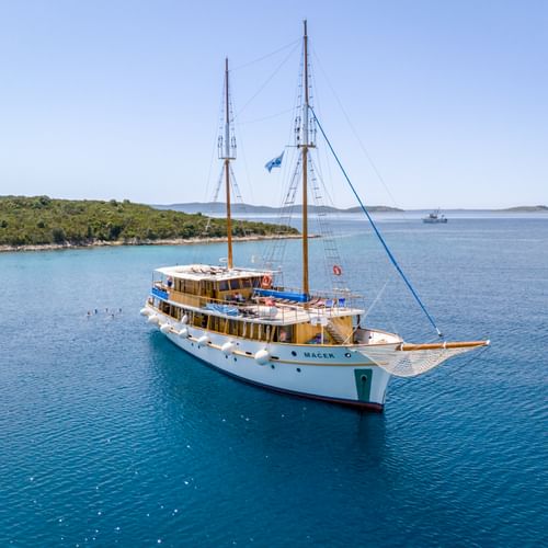 White motor sailing yacht MS Macek with two masts anchored in turquoise Adriatic Sea near green Croatian coastline under clear blue sky.