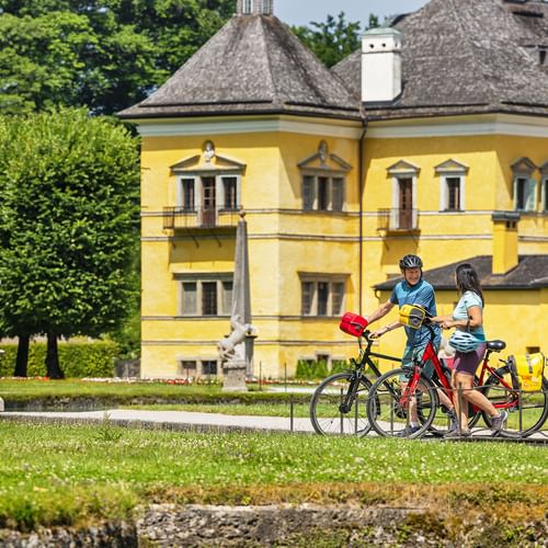 Zwei Radfahrer mit Fahrrädern auf einer Wiese vor dem gelben Schloss Hellbrunn in Salzburg, mit Statuen und grünen Bäumen im Park.