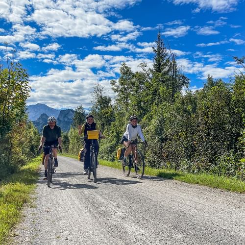 Three cyclists riding on a gravel path along the Salzach cycle route near Rif, surrounded by green trees with mountains in the background.