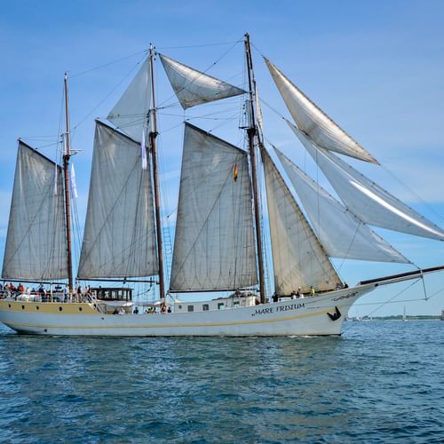 Three-masted sailing ship Mare Frisium with white sails unfurled on calm blue water under a clear sky. Dutch flag visible at stern.